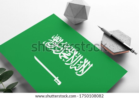 Saudi Arabia flag on minimalist paper background. National invitation letter with stylish pen on stone. Communication concept.