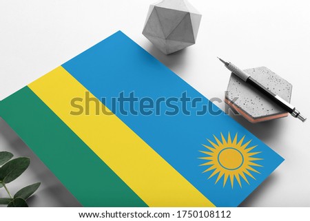 Rwanda flag on minimalist paper background. National invitation letter with stylish pen on stone. Communication concept.