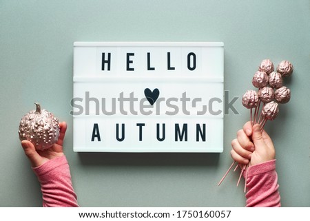Lightbox with text Hello Autumn and heart on silver grey paper background. Flat lay with female hands holding pink metallic pumpkin and small apples.