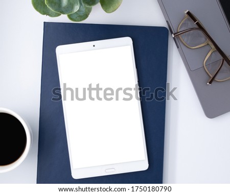 White office desk table with blank notebook, white blank mockup screen Tablet, laptop compute, glasses, cup of black coffee and supplies. Top view with copy space, flat lay.
