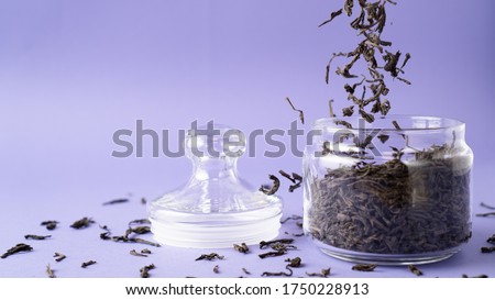 Large leaf black tea in glass jar. Flying tea leaves. Falling dried green tea leaves into bottle. Purple background. Royalty-Free Stock Photo #1750228913