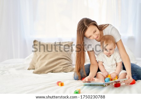 A young, beautiful mother with long hair sitting on the bed with her baby leafs through a children's book.