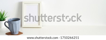 White desk with a coffee mug and a mockup frame. Blank wall copy space. Panorama