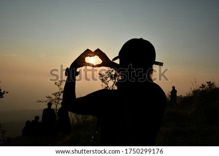 The black silhouette of a young traveler makes a heart shaped hand symbol with the light of the morning sun shining, to tell love