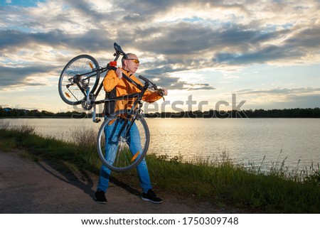 Tourist biker carries a bicycle on his shoulder against the backdrop of the setting sun on the lake.