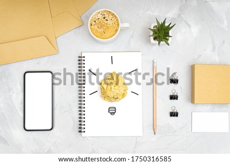 Refreshment break idea and productivity boost concept. Flat lay of an office workplace with a smartphone, coffee cup, light bulb symbol made of a crumpled paper ball and a sketch on a spiral notebook.