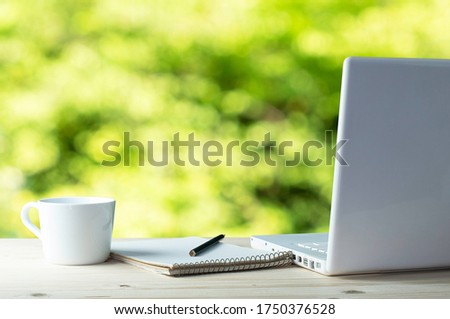 Workplace with white laptop, notebook and coffee on wooden desk ,which has a backdrop in a blurred natural green garden. Royalty-Free Stock Photo #1750376528