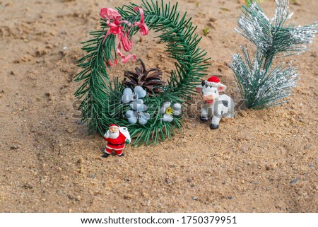 Year of the bull. Year of the cow. New year 2021. Cow in a Christmas hat. Santa Claus with a bag of gifts. Spruce with a cone. Objects stand on the sand.