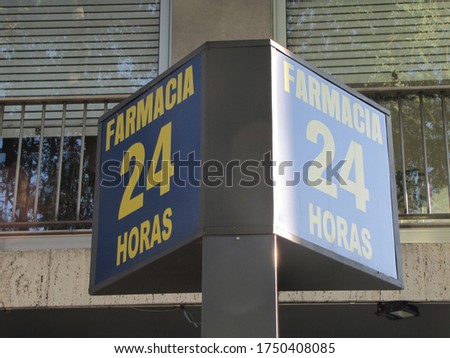 Poster with the sign, translation: "24 hour pharmacy" on the sidewalk. Street sign in blue with letters and yellow cross with sunlight illuminated apartment building background.