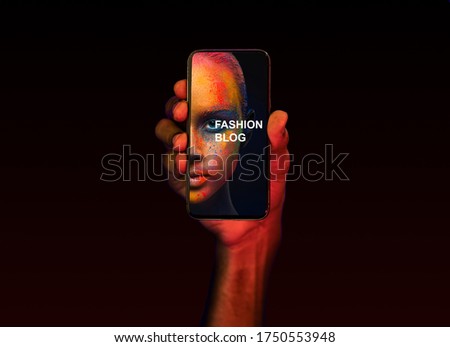 Male hand holding smartphone with FASHION BLOG webpage on screen against black background, creative collage