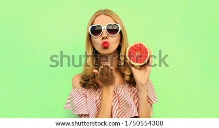 Summer portrait of beautiful young woman with slice of juicy grapefruit blowing her lips sending sweet air kiss wearing heart shaped sunglasses on green background