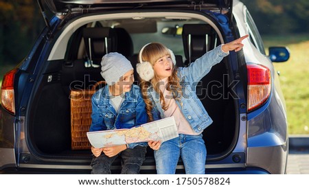 Little cute girl shows something to her brother while they are sitting in the opened car's trunk and looking to the road map. Royalty-Free Stock Photo #1750578824