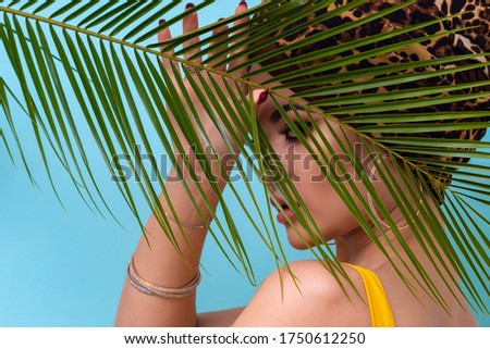An attractive young woman in a stylish turban made of leopard print fabric on a blue background. Girl with bright makeup. Beach style, palm branch and heat