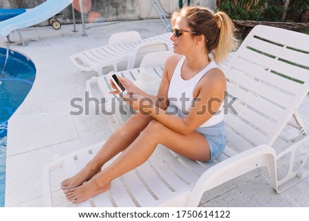 Stock photo of a caucasian woman sitting on the poolside. She is using her mobile phone. She is wearing sunglasses.