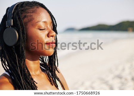 Face of beautiful young Black woman smiling and closing eyes when listening to relaxing music in her headphones and enjoying sea breeze