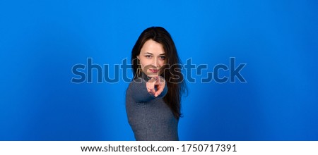 Photo of smiling, cheerful young female in grey dress on blue background. Emotional portrait