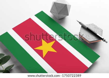 Suriname flag on minimalist paper background. National invitation letter with stylish pen on stone. Communication concept.