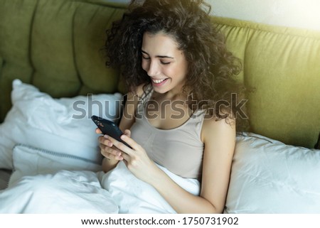 Above top view head shot joyful beautiful brunette woman relaxing under duvet in bed, using mobile applications. Smiling pleasant young lady typing message for friends in smartphone after wake up.