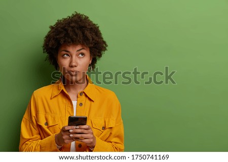 Pensive woman uses modern application on smartphone, thinks about message content, concentrated above, stays in touch with modern technologies, dressed in fashionable clothes blank space on green wall