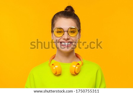 Young happy teenage girl wearing neon green sweatshirt, eyeglasses and headphones around neck, smiling and laughing, isolated on yellow background