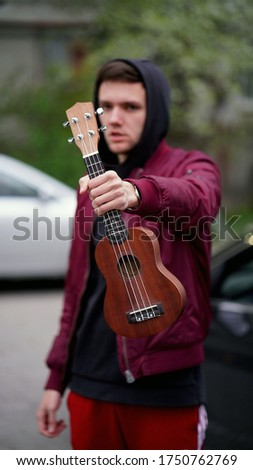Young unshaven man in a red jacket with a black hood with a ukulele in his hand outdoors