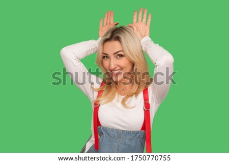 Portrait of carefree beautiful adult blond woman in stylish denim overalls showing bunny ears and smiling with happy childish playful expression. indoor studio shot isolated on green background