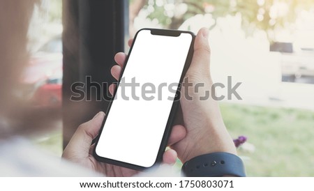 Mockup image of hand holding black mobile phone with blank white screen on thigh with white canvas shoes at vintage tile floor in cafe , feeling relax
