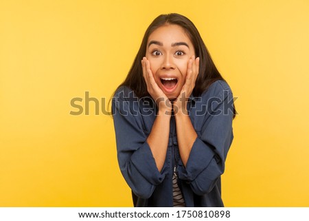 Oh my god! Portrait of excited shocked girl in denim shirt standing, grabbing her face and shouting in amazement, surprised by unbelievable success. indoor studio shot isolated on yellow background