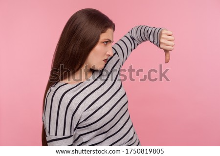 Dislike! Side view of angry dissatisfied woman in striped sweatshirt showing thumbs down and frowning displeased, expressing disapproval, awful feedback. indoor studio shot isolated on pink background