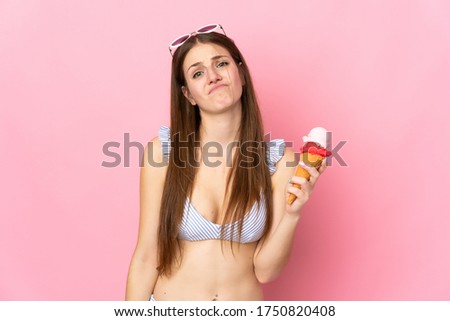 Young caucasian woman in swimsuit holding a cornet ice cream isolated on pink background with sad expression