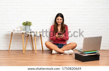 Young woman with a laptop sitting on the floor at indoors sending a message with the mobile