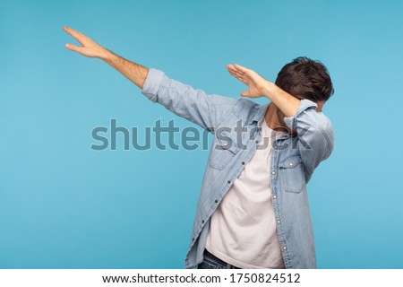 Dab dance. Portrait of man in denim shirt making dabbing movement, famous internet meme of success victory, expressing happiness and following trends. indoor studio shot isolated on blue background Royalty-Free Stock Photo #1750824512