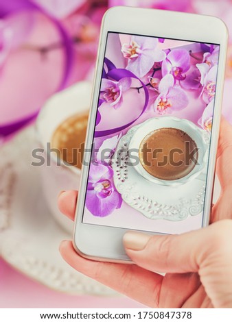 Woman taking beautiful picture of cup of coffe with pink orchids and purple ribbons on table with her smartphone. Close up