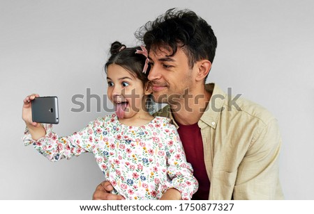 Playful little daughter and her smiling father  showing tongue and making grimace while taking self portrait on smart phone at home. Dad and cute little girl taking a selfie on mobile phone.