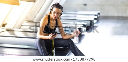 Woman athlete taking rest and listen to music on mobile phone after running on treadmill machine at gym sports club. Fitness Healthy lifestye and workout at gym concept.
