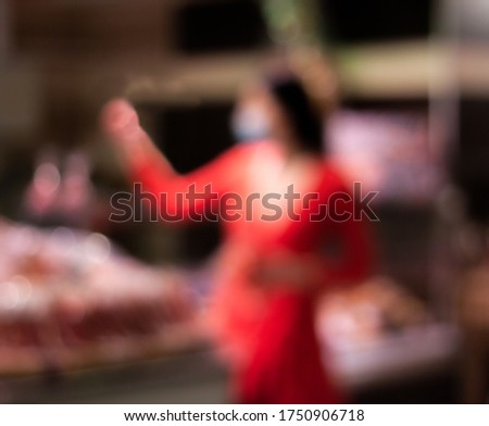  Beautiful  brunette girl picking up a pack of sliced meat in the supermarket. She is dressed in a bright red dress and medical protective  mask, the picture is totally blurred 