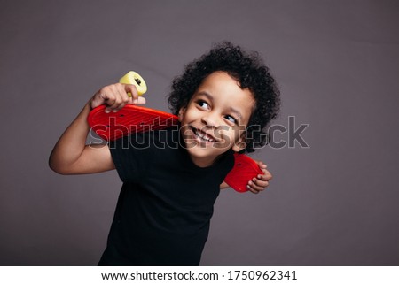 Close-up portrait of a curly African-American boy in black T-shirt holding a red skateboard behind his back and smiling at camera.