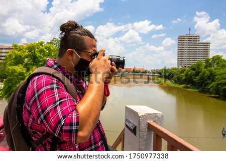 Young male asian traveler backpacker in hipster hair style with camera and backpack take a pictures of Chiangmai scenery by the river. Travel and holiday concept.
