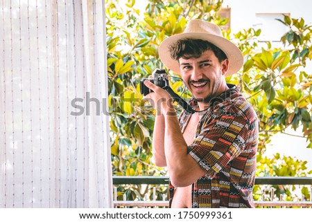 Attractive young man with a fashionable shirt looks at camera from the balcony. Laughs happily as he shoots a picture on his vacation.