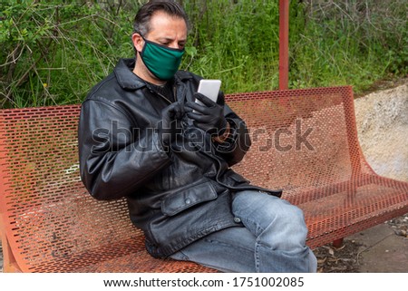 Man wearing mask and gloves sitting on a bench during covid 19 pandemic threat and looking his smartphone