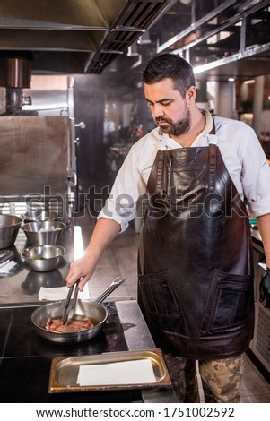 Busy portly chef with beard standing at stove and using tongs while frying steak