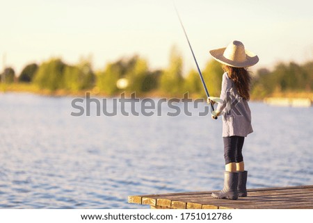 Cute little child girl in rubber boots and straw hat on wooden pier on a lake. Family leisure activity during summer sunny day.
