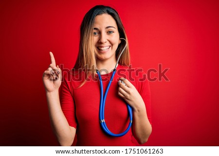 Young beautiful brunette woman using stethoscope standing over isolated red background surprised with an idea or question pointing finger with happy face, number one
