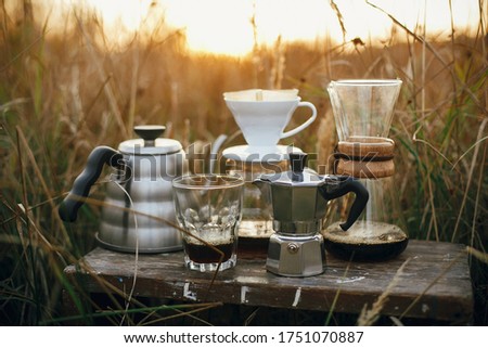 Alternative coffee brewing outdoors in travel. Steel kettle, hot coffee in cup, coffee dripper,  geyser maker, glass flask with filter on background of sunny warm light in rural herbs. Royalty-Free Stock Photo #1751070887