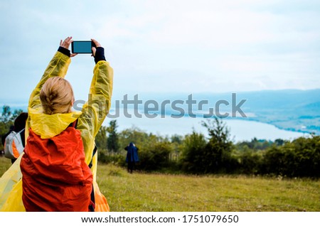 Girl hiker taking a photo of beautiful scenery in nature using mobile phone. Woman take a picture of river with phone on the hill, standing in rainy yellow coat with red backpack. Copy space