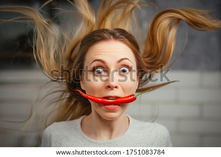 A girl with a red hot pepper in her teeth. Chili for cooking. The spicy seasoning is very stinging in the mouth. My hair stood on end. Bite the pepper and get burned. Emotional woman. Royalty-Free Stock Photo #1751083784