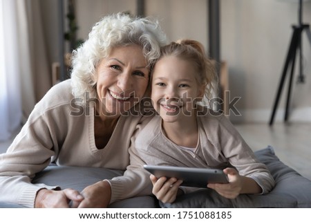 Portrait of smiling senior grandmother and little granddaughter lying on floor playing using tablet together, happy mature grandparent and small grandchild relax have fun with pad gadget at home