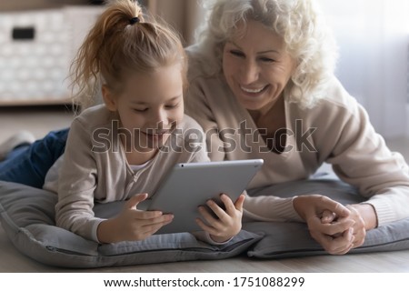 Playful happy elderly grandmother and little granddaughter relax lying on floor at home watching video pad gadget, smiling mature grandparent have fun play using modern tablet with small grandchild