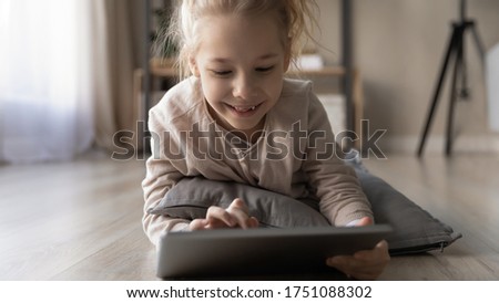 Smiling little preschooler girl child lying on pillow on home floor playing on tablet gadget, happy small kid relax rest in living room, have fun using application on pad device, technology concept