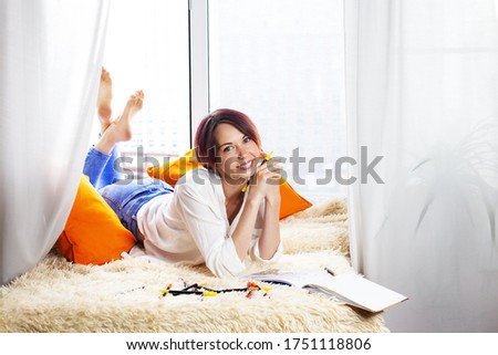 Cheerful young woman drawing with sketch markers in her sketcbook and lying near the window on yellow furry plaid with orange pillows. Interesting hobby at home, leisure during social distancing. 